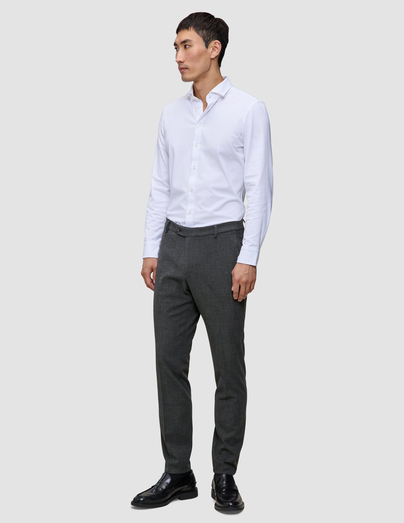 En mand iført en hvid skjorte med knapper, Motion Suit-bukser i Slim-pasform i farven Pepper Grey og sorte lædersko.